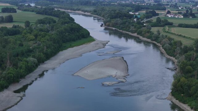 Dry sandbar and mudflat emerge from the Po River amid low water levels, with farmland and rural buildings along the banks in the Po Valley countryside, drone ascent.