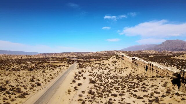 Lonely paved highway runs past a rugged desert cliff and scrubland in Catamarca under clear blue skies, with wide drone flyover over arid terrain and distant mountains.