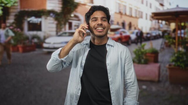 Young man holds smartphone to ear smiling on street, gestures with hands while speaking into device in busy urban lane; joy connection.