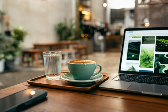 Content creator working on laptop in cafe. Browsing inspiration photos with coffee and smartphone on desk.