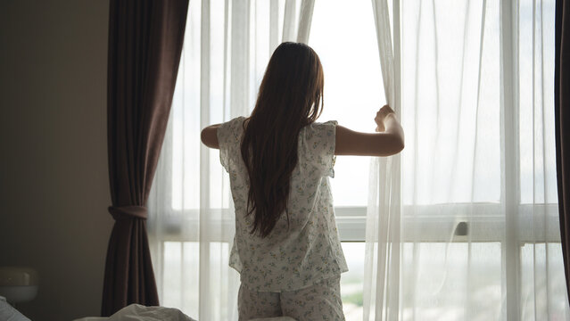 Rear view of young woman opening curtains in bedroom in the morning