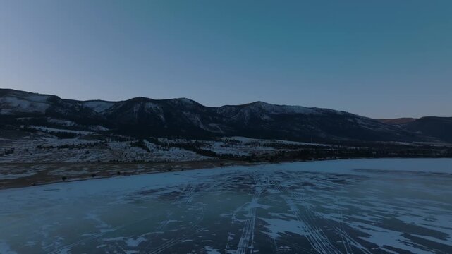 Drone footage over frozen Lake Baikal with cracked blue ice and snow patterns, surrounded by mountains and shoreline in Russia, winter landscape background