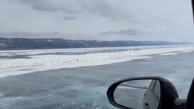 POV from a car on the frozen surface of Lake Baikal, showing an ice road, snow ridges, and distant mountains under an overcast sky in Siberia, Russia
