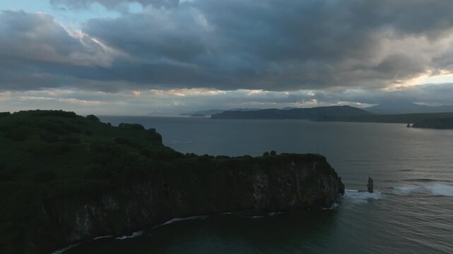 Drone footage of rugged coastal cliffs and the Chertov Palets sea stack at Bolshaya Lagernaya Bay, Kamchatka, Russia, under dramatic clouds above the ocean
