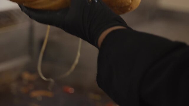 Chef filling a long bread roll with hot chicken, grilled vegetables and melted cheese, preparing a loaded takeaway sandwich in a restaurant.