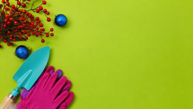 Gardening tools including a turquoise trowel and pink gloves arranged with Christmas decorations like red berries and blue ornaments on a vibrant green background