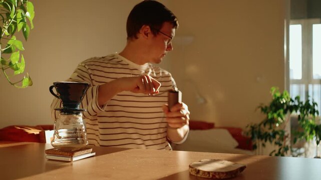 Young man using a manual burr grinder to prepare freshly roasted coffee beans for a pour over brew, enjoying his morning routine in a warmly lit kitchen with plants
