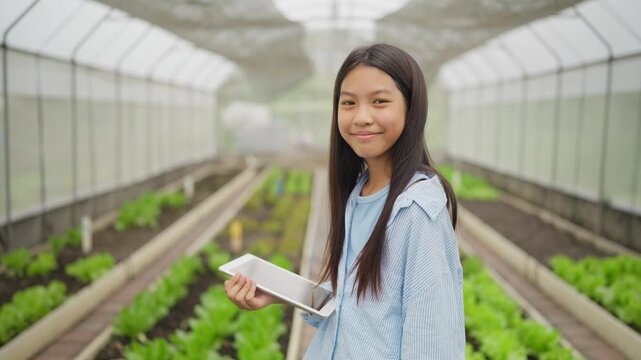 Young woman smiles in greenhouse holding tablet. She walks among rows of lettuce plants. Rows stretch long under bright plastic roof