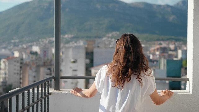 Young woman enjoying a scenic city and mountain view from a high-rise balcony on a bright sunny summer day, relaxing and appreciating the peaceful urban landscape.
