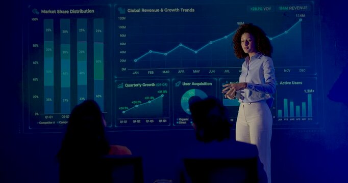 Adult female business presenter speaks beside digital performance dashboard, using expressive gestures while explaining growth analytics to seated audience during corporate intelligence presentation.