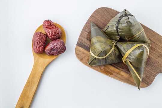 Triangular zongzi wrapped in leaves with red dates on a wooden spoon, placed on a wooden board against a white background.