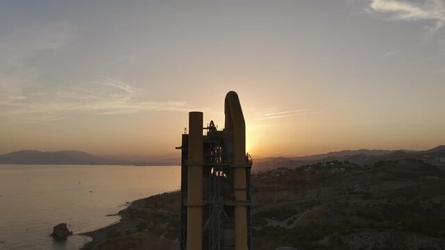Silhouette of an industrial cement factory tower against a dramatic sunset sky along the coast near Malaga, Spain.
