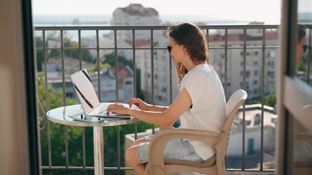 Young woman working on a laptop while sitting on a high-rise balcony overlooking the city on a sunny summer day, combining remote work with a relaxed outdoor lifestyle.