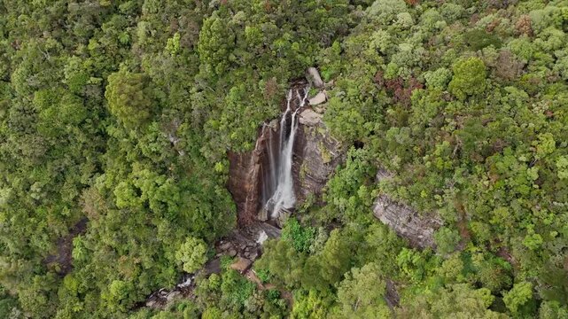 Cinematic aerial drone shot pulling backward and ascending over the cascading water of Lovers Leap waterfall revealing dense green jungle in Nuwara Eliya Sri Lanka