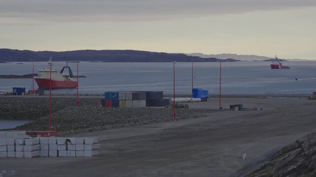 Telephoto tracking view of red ship Mersey Venture manoeuvring past icebreaker Captain Molly Kool heading toward artificial breakwater causeway at Iqaluit Deep Sea Port