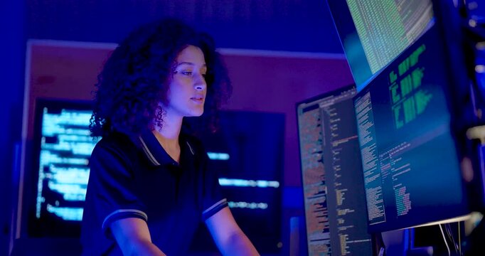 Adult female technology analyst watches multiple monitors, shifts attention between code displays, and continues review during cybersecurity monitoring and secure digital data operations at work.