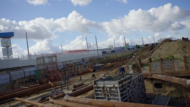 Worker operates equipment in deep excavation pit surrounded by support pipes. Sunny sky overhead highlights major urban foundation project