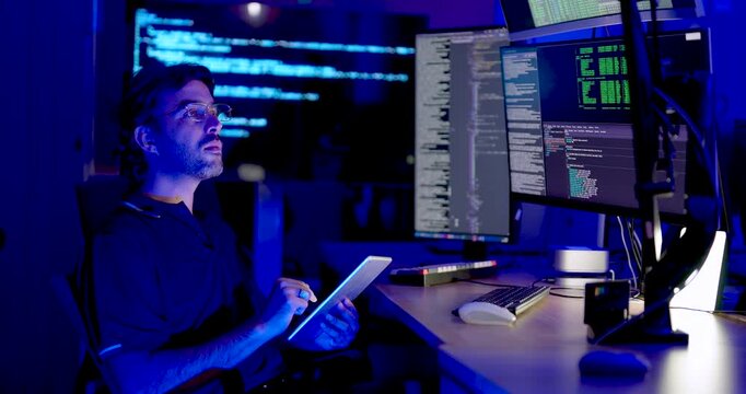 Adult Asian male cybersecurity analyst holds tablet, reviews information, and looks toward code monitors during digital system monitoring, network security analysis, and secure data operations.