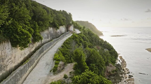 Winding coastal road carved into a lush green cliffside, overlooking the vast expanse of the ocean and Green Bowl Beach during a soft evening light in Bali island, Indonesia. Aerial drone flight
