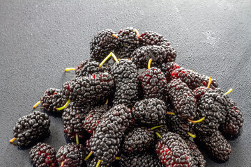 A pile of fresh, ripe dark mulberries rests on a dark textured surface, showcasing their bumpy, richly colored texture. © tai