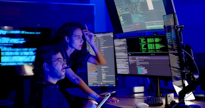 Adult male cybersecurity analyst reviews tablet beside female colleague as both watch code monitors, exchange gestures, and discuss system information during collaborative network security analysis.