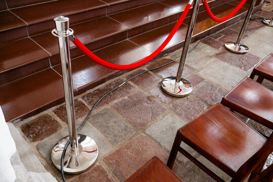 A detailed commercial shot of silver metallic crowd control barriers with luxury red velvet ropes standing on a stone floor next to wooden chairs, highlighting event organization and safety queue mana
