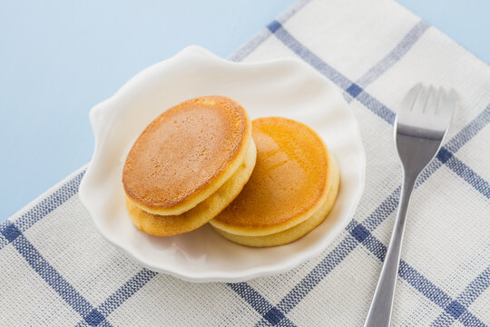 Several small fluffy pancakes are stacked on a white plate, placed on a checkered cloth with a fork beside.
