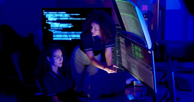Adult female cybersecurity analyst sits while female colleague leans closer, gestures toward laptop and code monitors, and collaborates on digital system review and network security analysis.