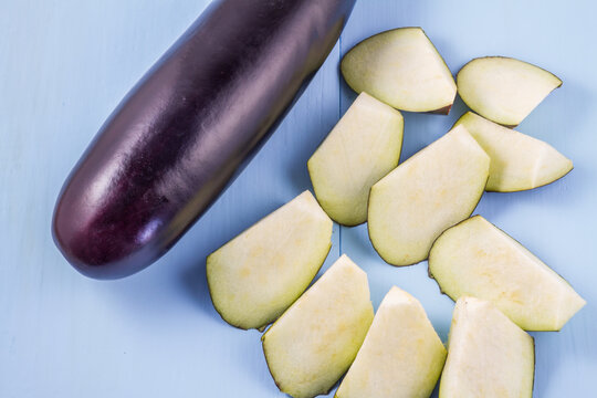 A whole raw eggplant and cut eggplant slices rest on a light blue flat surface, showing fresh vegetable produce.