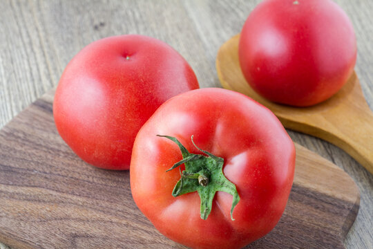 Several fresh ripe red tomatoes rest on dark wooden cutting board and light wooden spoon, placed on a light wood surface, ready for culinary use.