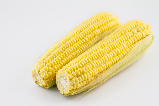 Two fresh raw corn cobs with bright yellow kernels, partially covered with light husks, placed on a plain white background.