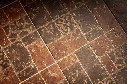 Close-up of rustic brown ceramic floor tiles featuring decorative patterns, visible grout lines and worn surface texture. Ideal for interior design, flooring projects, backgrounds and architecture