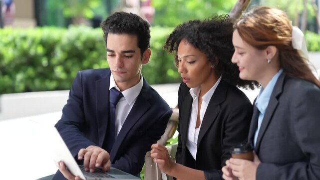 Three colleagues cluster around one laptop screen outdoors, smiling as a result comes in, a candid teamwork moment on a landscaped corporate plaza in the morning sun