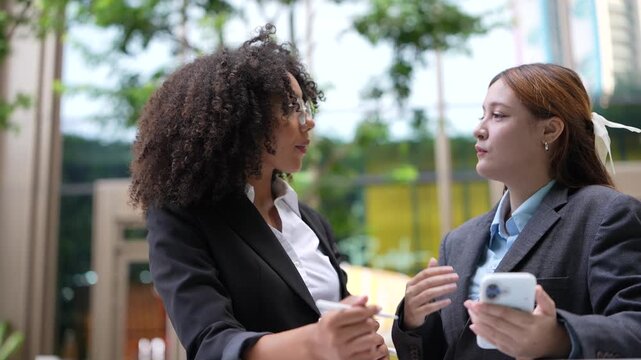 Businesswoman shows something on her smartphone to a colleague during an outdoor break, the two comparing screens beside a planted courtyard on a bright Bangkok morning