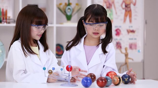 Two Young Girls in Lab Coats Studying Solar System Model in Classroom