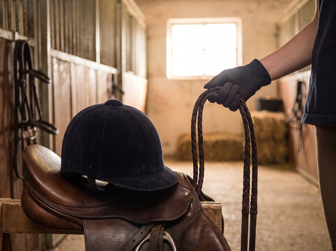 Equestrian rider holding reins in stable with riding gear beside  
