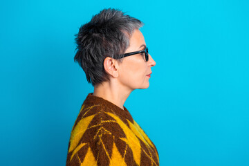 Profile of a mature woman with short gray hair and glasses against a vibrant blue background wearing a patterned yellow and brown sweater © deagreez
