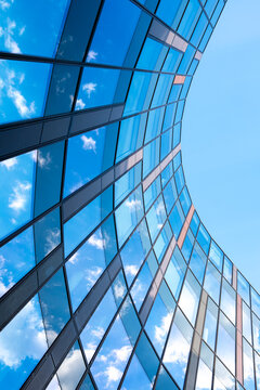 Curved blue glass facade reflects clouds and sky, creating a dynamic low angle view of contemporary architecture with sweeping lines and mirrored surfaces.