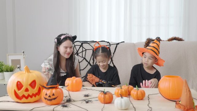 Asian family enjoys fun Halloween celebration at home, young siblings in costumes, mother showing jar, decorative pumpkins and spiders adorn table, creating festive bonding time.