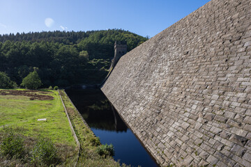 Low-angle view of the massive stone masonry wall and Gothic turret tower of Derwent Dam rising against forested hills and a blue sky in Derbyshire, England © bardhok