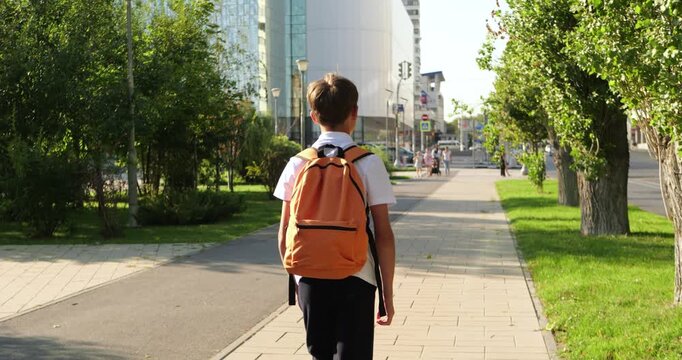 Schoolboy walks through city park with orange backpack. Young student heads to school on sunny urban sidewalk. Green trees and modern buildings frame independent back to school journey.