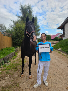 Woman holds certificate next to horse on a dirt path with trees
