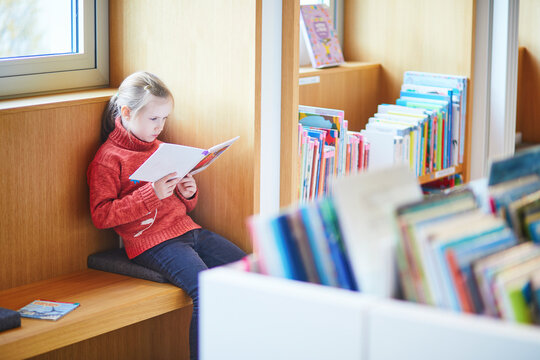 Girl reading a children's book in a French city library, sitting by a window among bookshelves on a quiet winter day