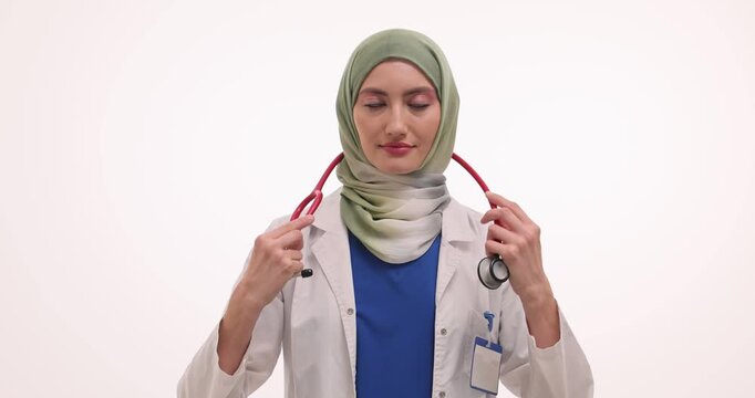Smiling woman doctor in hijab places stethoscope around neck. Muslim physician prepares for thorough examination of patient against white background