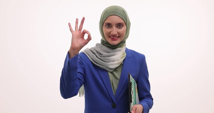 Businesswoman in hijab holds detailed statistical report in hand. Successful Muslim executive confidently shows OK gesture at white office wall