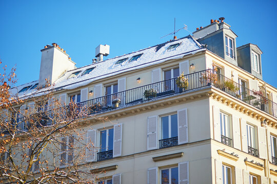 Parisian apartment building with a snow-covered mansard roof, white shutters and balconies under a bright blue winter sky