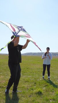 Young caucasian couple unsuccessfully attempting to launch a colorful kite on a windy day, laughing together in a vast green meadow under a clear blue sky during their fun outdoor activity