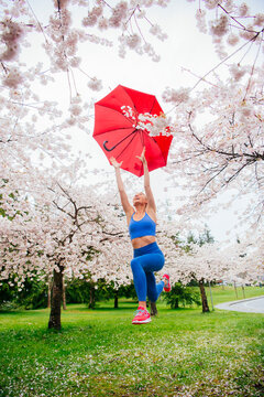 White woman jumping under cherry blossom trees with red umbrella