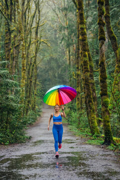 White woman running on rainy forest trail holding rainbow umbrella