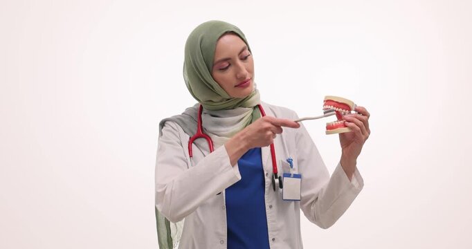 Woman dentist in hijab shows correct brushing technique. Doctor uses artificial jaw model to show proper brushing movements against white background